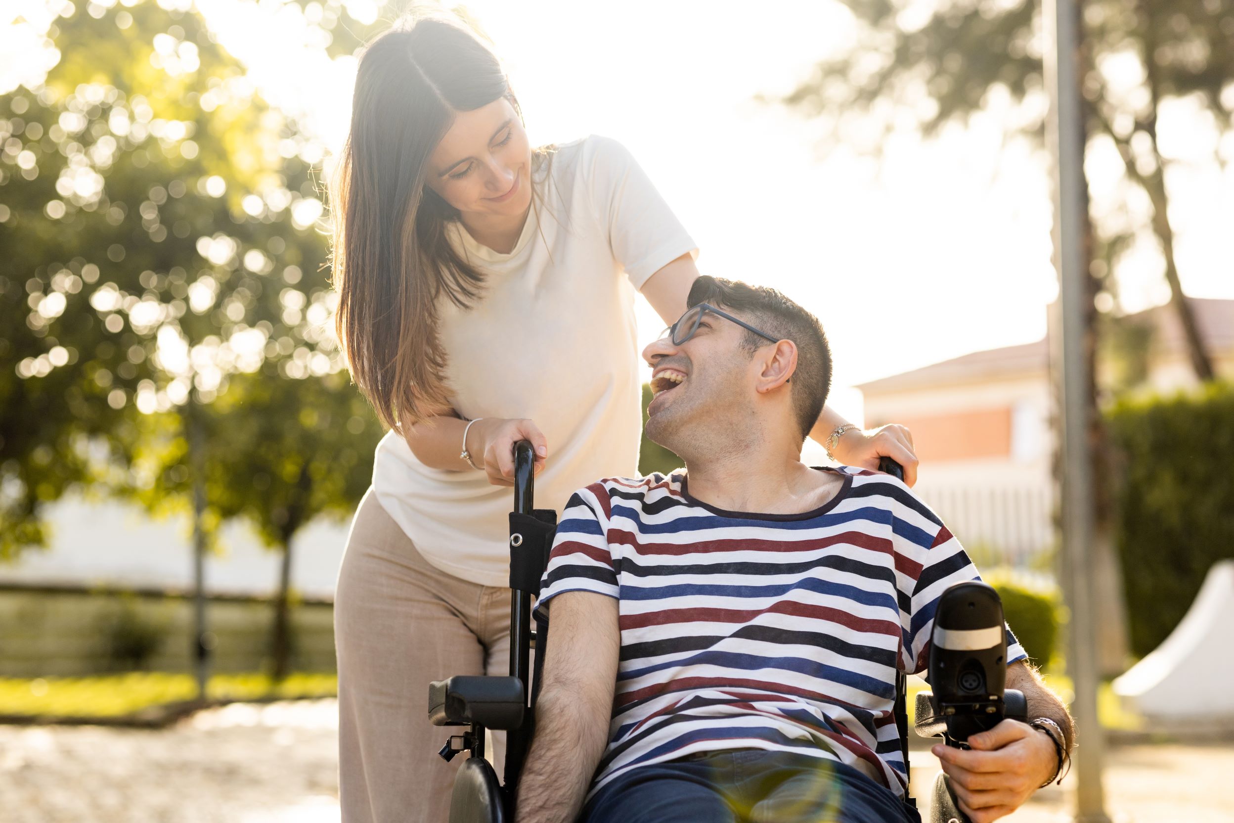 Woman stands in park outdoors looking fondly at smiling man next to her who has a disability and uses motorized wheelchair.